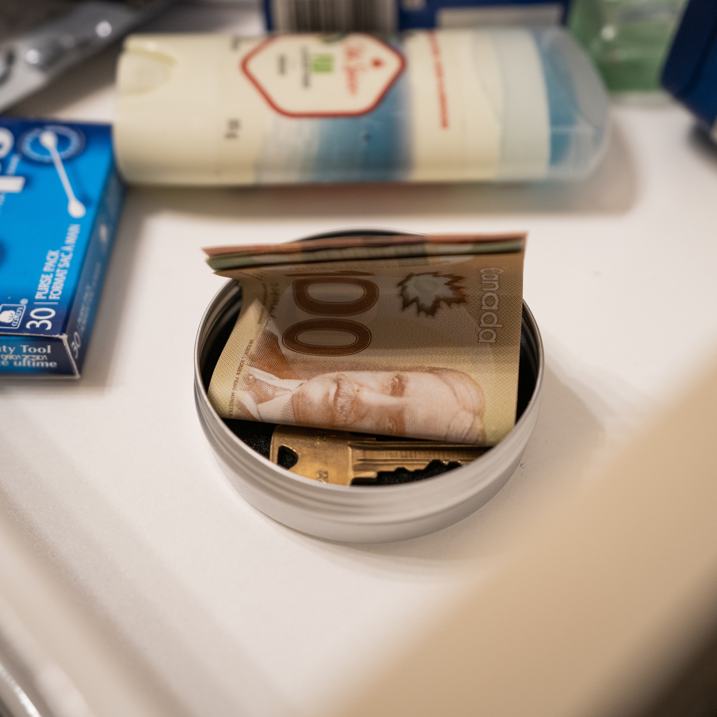 Compact metal tin storing Canadian $100 bill and keys, placed discreetly on a bathroom counter among everyday items.